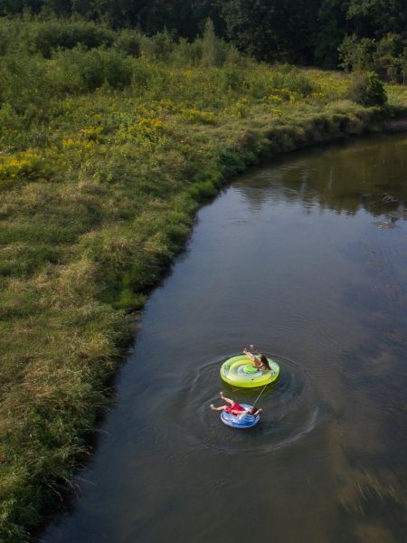 Two people float on inflatables down a winding river surrounded by lush green trees and grass near Village Green apartments.