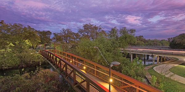 A scenic view of a lit-up wooden bridge amidst trees under a colorful sunset sky with a visible moon near Village Green Apartments.