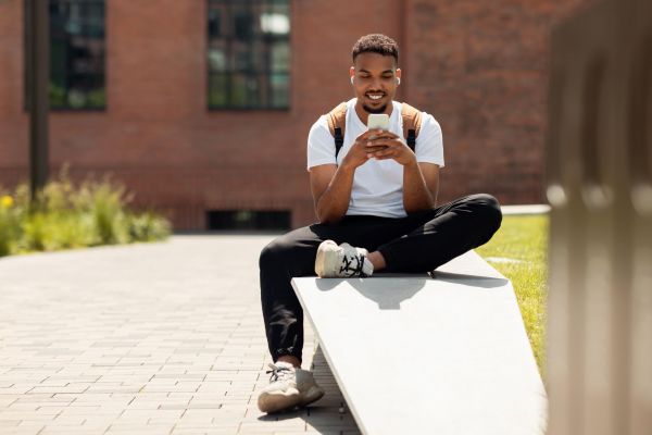 Village Green apartments resident sitting outside on a bench, smiling while looking at his phone, wearing a backpack and earphones.