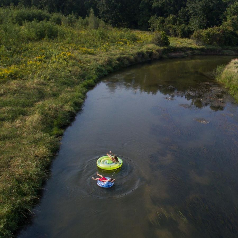 Two people float on inflatables down a winding river surrounded by lush green trees and grass near Village Green apartments.