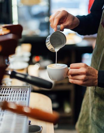 Barista pouring milk into a cup of coffee, creating latte art, while wearing a green apron at a cafe counter near Village Green apartments.