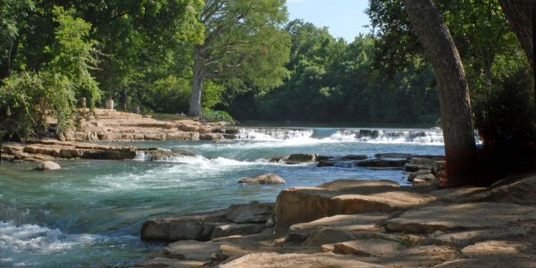 A serene river with gentle rapids flows through a lush, green forest next to rocky banks under a clear sky near Village Green Apartments.