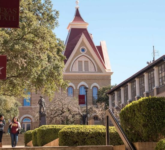 Two people walk on a campus path lined with trees, leading to a historic building under a clear blue sky.