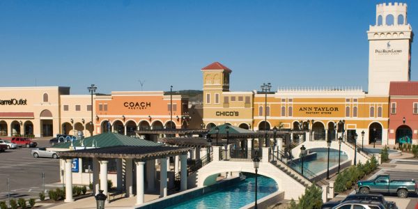 Outlet mall with water fountain, parking lot, and brand storefronts under a clear sky near Village Green Apartments.